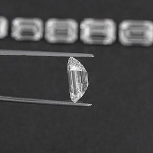 May include: A clear, faceted, emerald-cut gemstone held by tweezers against a black background.
