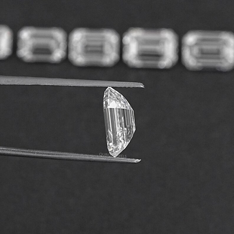 May include: A clear, faceted, emerald-cut gemstone held by tweezers against a black background.