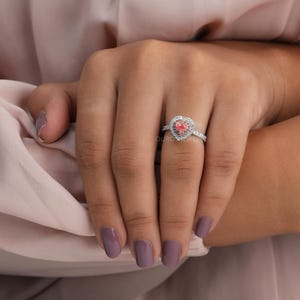 May include: A silver ring featuring a central heart-shaped pink gemstone surrounded by a halo of small white stones. The band is also encrusted with small white stones along the visible portion. The text OURO JEWELS is visible on the ring.