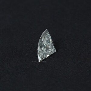 Puede incluir: Un diamante incoloro suelto con corte de escudo y una superficie facetada que refleja la luz. La piedra preciosa presenta una parte superior puntiaguda y un lado curvo, mostrando gran claridad y brillo. El texto OUROSJEWELS est&aacute; impreso en peque&ntilde;as letras blancas.