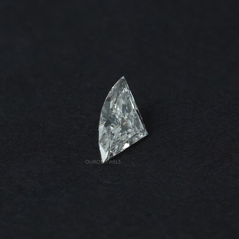 Puede incluir: Un diamante incoloro suelto con corte de escudo y una superficie facetada que refleja la luz. La piedra preciosa presenta una parte superior puntiaguda y un lado curvo, mostrando gran claridad y brillo. El texto OUROSJEWELS est&aacute; impreso en peque&ntilde;as letras blancas.