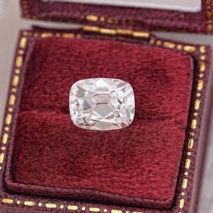 May include: A large, clear, cushion-cut diamond with many facets, set against a deep red velvet jewelry box. The diamond reflects light, showcasing its brilliance. The box has a gold-colored trim.