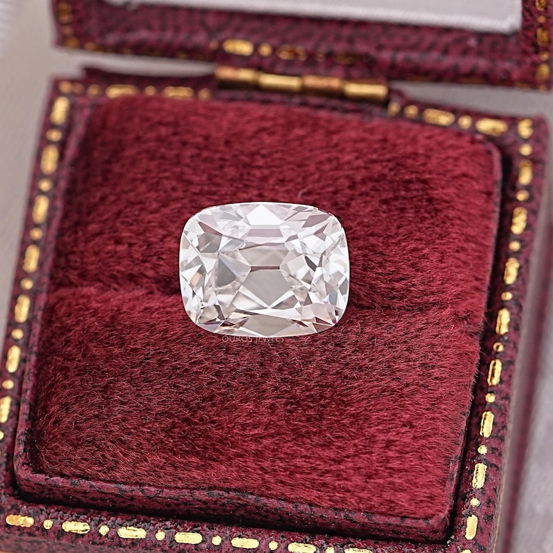 May include: A large, clear, cushion-cut diamond with many facets, set against a deep red velvet jewelry box. The diamond reflects light, showcasing its brilliance. The box has a gold-colored trim.