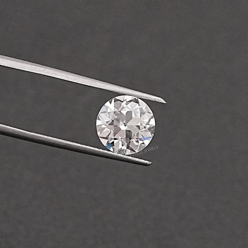 May include: A clear round gemstone with facets, held by tweezers against a gray background.