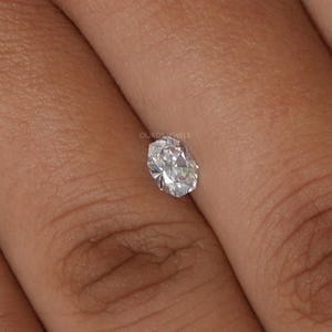 May include: A clear, oval-cut diamond with multiple facets, reflecting light. The diamond is held between fingers, showcasing its brilliance. The image is a close-up, highlighting the gem's clarity and shape. The text "OUROS JEWELS" is visible.
