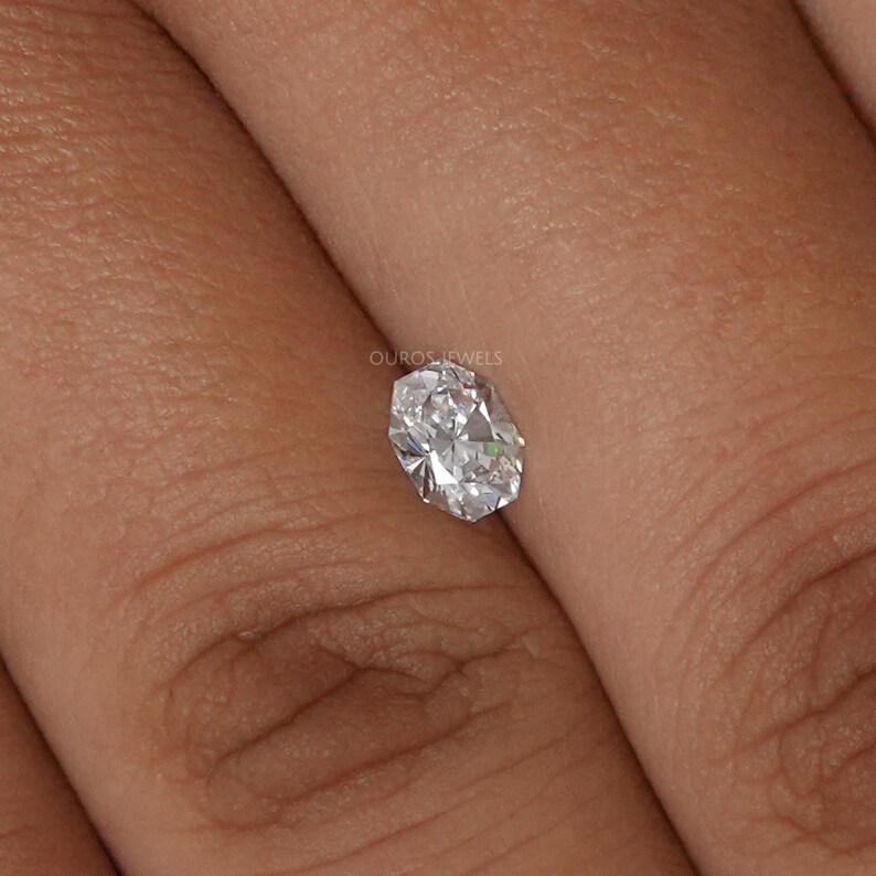 May include: A clear, oval-cut diamond with multiple facets, reflecting light. The diamond is held between fingers, showcasing its brilliance. The image is a close-up, highlighting the gem's clarity and shape. The text "OUROS JEWELS" is visible.