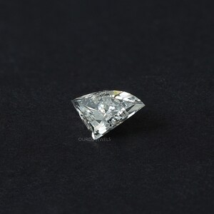 Puede incluir: Una piedra preciosa incolora de corte trillion con forma triangular y m&uacute;ltiples facetas que reflejan la luz. La piedra presenta una punta afilada y bordes curvos, con el texto OUROSJEWELS impreso en peque&ntilde;as letras blancas cerca de la base.