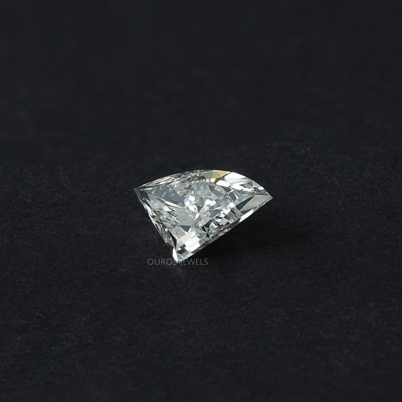 Puede incluir: Una piedra preciosa incolora de corte trillion con forma triangular y m&uacute;ltiples facetas que reflejan la luz. La piedra presenta una punta afilada y bordes curvos, con el texto OUROSJEWELS impreso en peque&ntilde;as letras blancas cerca de la base.
