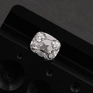 May include: A clear, cushion-cut diamond with numerous facets, reflecting light. The diamond is set against a black background. The text "OUROSJEWELS" is visible on the lower right side of the image. The diamond's shape is rectangular with rounded corners.
