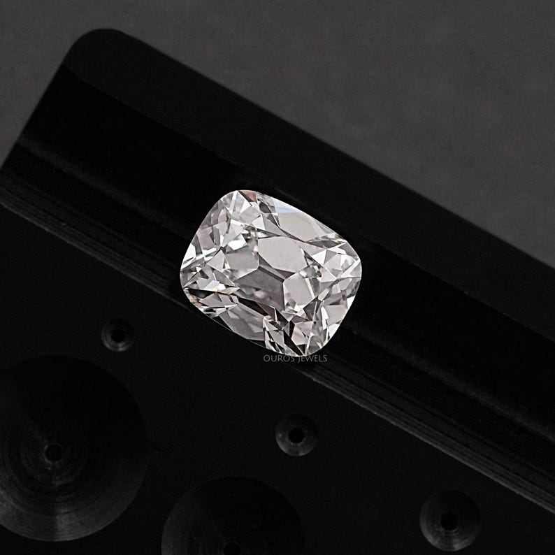 May include: A clear, cushion-cut diamond with numerous facets, reflecting light. The diamond is set against a black background. The text "OUROSJEWELS" is visible on the lower right side of the image. The diamond's shape is rectangular with rounded corners.