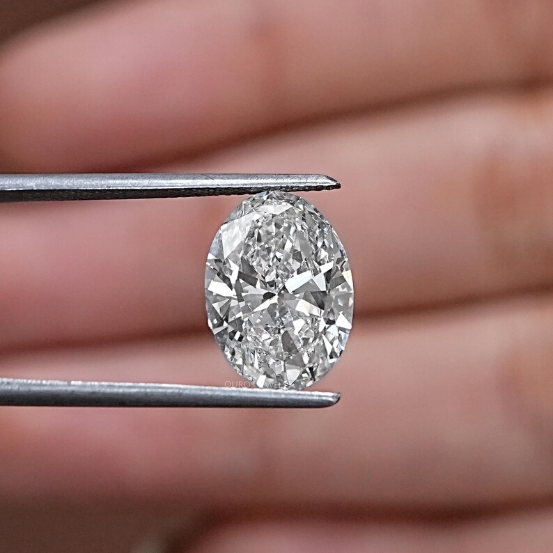 Pu&ograve; includere: Un diamante trasparente di forma ovale, tenuto da una pinzetta. Il diamante &egrave; sfaccettato e riflette la luce.