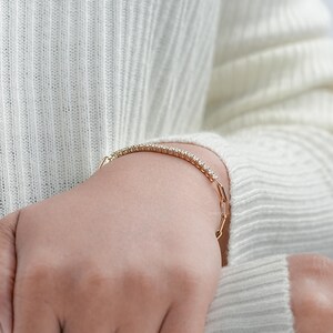 Pu&ograve; includere: Un braccialetto d'oro con una fila di piccole pietre preziose trasparenti. Il braccialetto &egrave; su un polso e la persona indossa un maglione bianco. Il braccialetto ha un design delicato ed elegante.