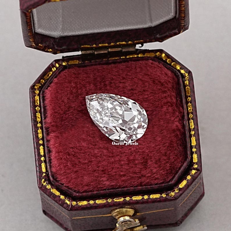 May include: A pear-shaped diamond gemstone in a red velvet jewelry box. The box has a gold trim and the text "Ouros Jewels" is printed on the bottom.