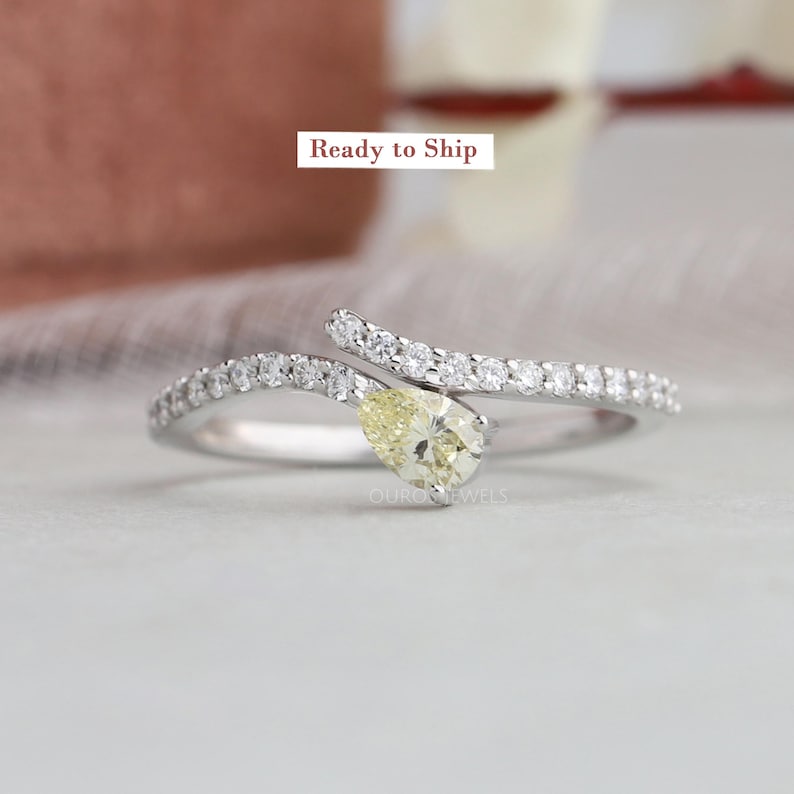 Peut inclure: Une bague bypass argent&eacute;e avec une pierre centrale jaune p&acirc;le en forme de poire et un anneau serti de petites pierres claires. La bague est plac&eacute;e sous une &eacute;tiquette rectangulaire portant le texte Ready to Ship et la marque Ourojewels.