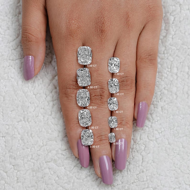 K&ouml;nnte beinhalten: Ein Handmodell mit helllila lackierten N&auml;geln zeigt einen Gr&ouml;&szlig;envergleich von Kissen-Schliff-Diamanten. Die Diamanten reichen von 0,50 Karat bis 5,00 Karat. Jeder Diamant ist mit seinem Karatgewicht gekennzeichnet.