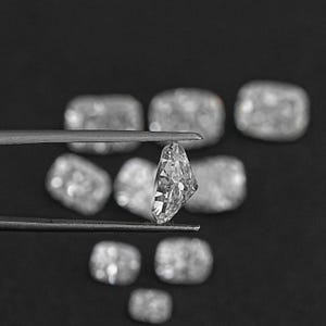 K&ouml;nnte beinhalten: Ein klarer, facettierter, rechteckiger Diamant-Edelstein, der mit einer Pinzette gehalten wird. Der Diamant ist von anderen &auml;hnlichen Edelsteinen auf schwarzem Hintergrund umgeben.