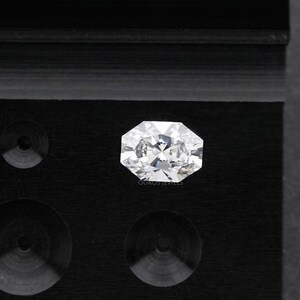May include: A brilliant-cut, octagonal-shaped diamond, set against a black background. The diamond sparkles with numerous facets, reflecting light. The image is a close-up, highlighting the clarity and brilliance of the gemstone. The text "OUROS JEWELS" is visible.