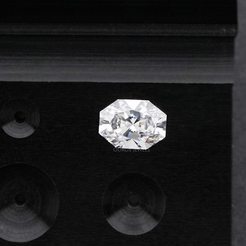 May include: A brilliant-cut, octagonal-shaped diamond, set against a black background. The diamond sparkles with numerous facets, reflecting light. The image is a close-up, highlighting the clarity and brilliance of the gemstone. The text "OUROS JEWELS" is visible.