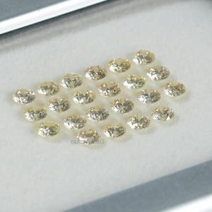 May include: A collection of 20 oval-shaped, light yellow gemstones. The gemstones are arranged in a semi-circle pattern on a white surface.