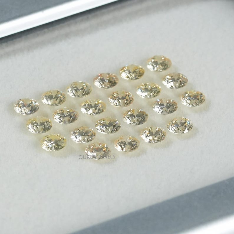 May include: A collection of 20 oval-shaped, light yellow gemstones. The gemstones are arranged in a semi-circle pattern on a white surface.