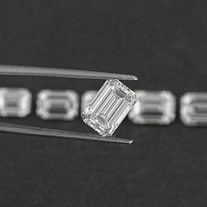 May include: A clear emerald cut diamond held by tweezers. The diamond is surrounded by other smaller diamonds.