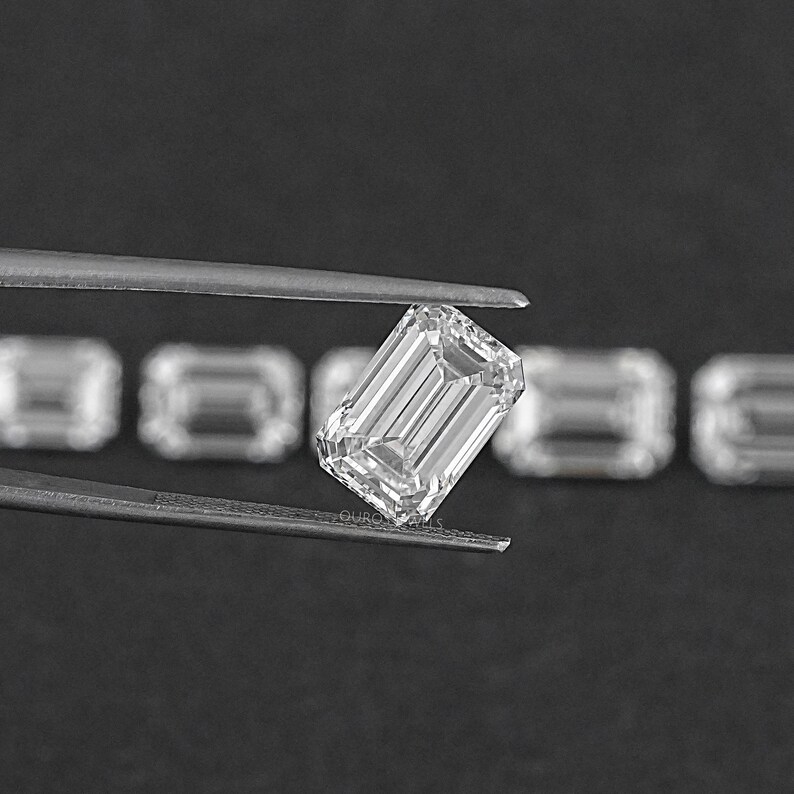 May include: A clear emerald cut diamond held by tweezers. The diamond is surrounded by other smaller diamonds.