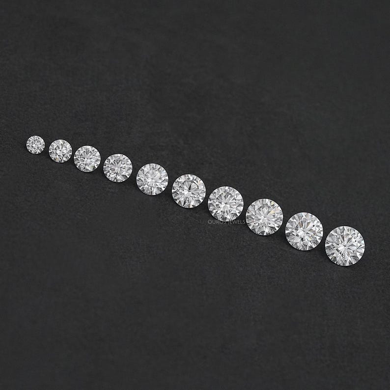 May include: A row of round, clear, faceted gemstones of varying sizes, arranged from smallest to largest on a black surface.
