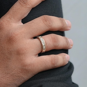 Puede incluir: Un anillo dorado con una serie de piedras preciosas claras de corte cuadrado engastadas alrededor de la banda. El anillo est&aacute; en un dedo, con la mano apoyada contra una tela oscura. El anillo tiene un dise&ntilde;o grabado en los bordes.