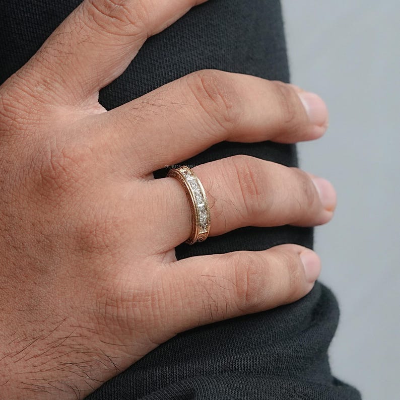 Puede incluir: Un anillo dorado con una serie de piedras preciosas claras de corte cuadrado engastadas alrededor de la banda. El anillo est&aacute; en un dedo, con la mano apoyada contra una tela oscura. El anillo tiene un dise&ntilde;o grabado en los bordes.