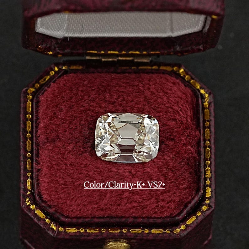 May include: A cushion-cut diamond with a light hue, set in a burgundy velvet jewelry box. The diamond's facets reflect light, creating a brilliant sparkle. Text on the box reads "Color/Clarity-K+ VS2+".