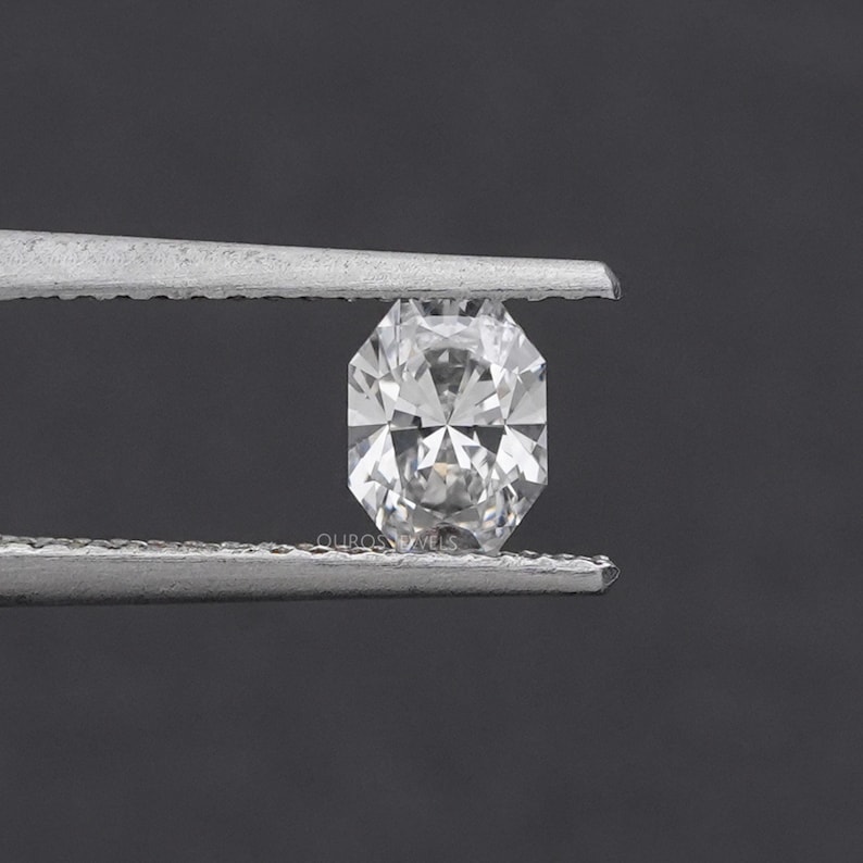 May include: A clear, faceted diamond held by silver tweezers. The diamond has an octagonal shape with multiple facets, reflecting light. The background is a dark gray, providing contrast to the bright gemstone. The text "OUROS JEWELS" is visible.