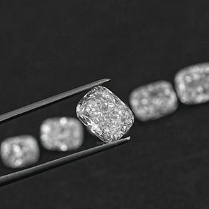 K&ouml;nnte beinhalten: Eine Reihe von klaren, facettierten, kissenf&ouml;rmigen Diamanten, die mit einer Pinzette gehalten werden. Die Diamanten sind in einer Linie angeordnet, wobei der gr&ouml;&szlig;te Diamant in der Mitte liegt.