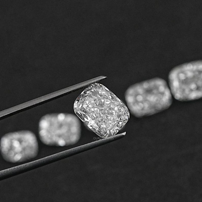K&ouml;nnte beinhalten: Eine Reihe von klaren, facettierten, kissenf&ouml;rmigen Diamanten, die mit einer Pinzette gehalten werden. Die Diamanten sind in einer Linie angeordnet, wobei der gr&ouml;&szlig;te Diamant in der Mitte liegt.