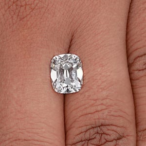 May include: A cushion-cut diamond with a brilliant, clear appearance. The gemstone is held in a hand, showcasing its facets and internal reflections. The diamond's shape is rectangular with rounded corners, and it sparkles with light.