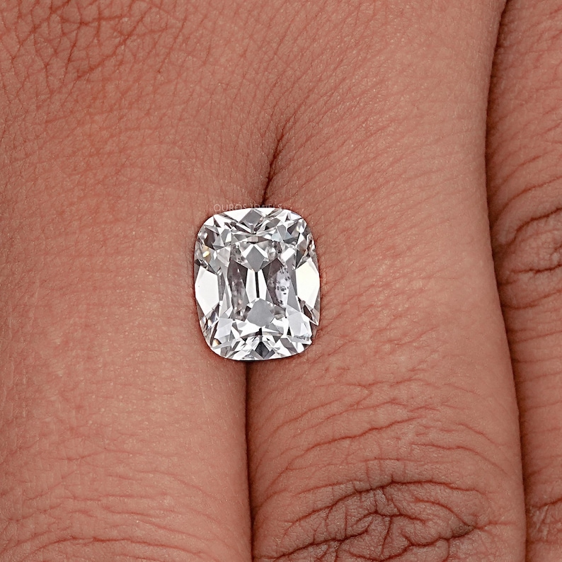 May include: A cushion-cut diamond with a brilliant, clear appearance. The gemstone is held in a hand, showcasing its facets and internal reflections. The diamond's shape is rectangular with rounded corners, and it sparkles with light.