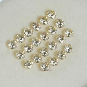 May include: A collection of 18 round, clear, faceted gemstones with a slight yellow hue. The gemstones are arranged in a semi-circle pattern on a white surface.