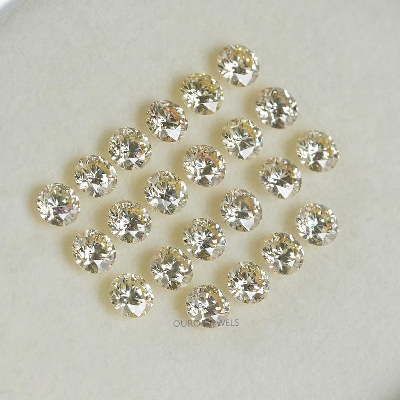 May include: A collection of 18 round, clear, faceted gemstones with a slight yellow hue. The gemstones are arranged in a semi-circle pattern on a white surface.