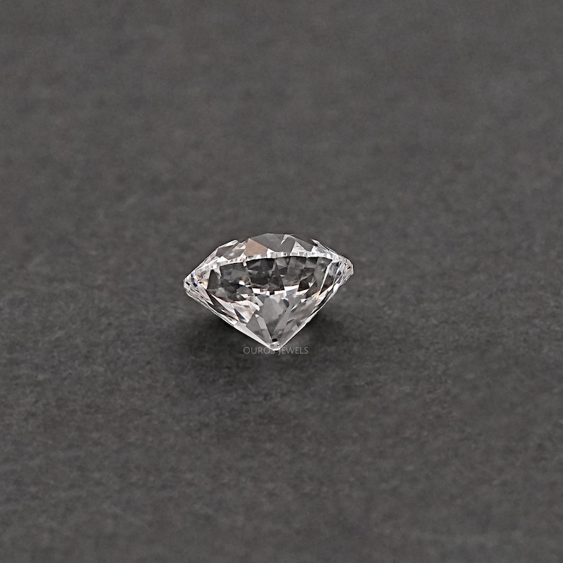 May include: A clear, round, faceted gemstone with a brilliant cut. The gemstone is set against a dark gray background.
