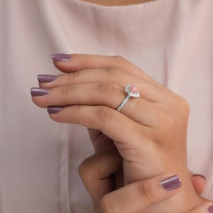 May include: A pear-shaped pink gemstone ring set in a silver-toned metal band. The central pink stone is surrounded by a halo of small white stones, with additional white stones embedded along the shank of the ring. The text OUROS JEWELS is visible.