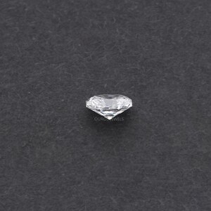 May include: A clear, round-cut diamond with multiple facets, reflecting light. The diamond is set against a dark gray background. The text "OURO JEWELS" is visible on the diamond.
