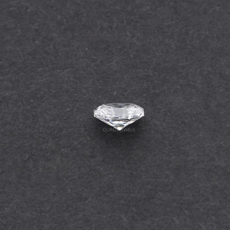 May include: A clear, round-cut diamond with multiple facets, reflecting light. The diamond is set against a dark gray background. The text "OURO JEWELS" is visible on the diamond.