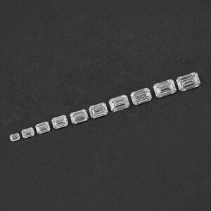 May include: A row of ten clear emerald cut diamonds of varying sizes, arranged in ascending order from left to right on a black surface.