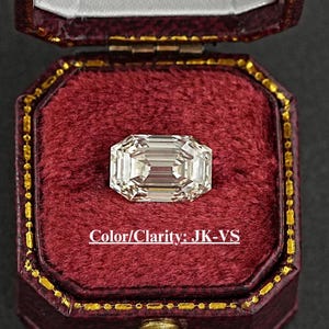 May include: A clear emerald cut diamond in a red velvet ring box. The diamond is in a setting with a gold band. The text "Color/Clarity: JK-VS" is printed on the box.