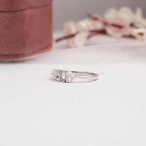 May include: A silver-toned metal ring featuring a central rectangular setting flanked by two smaller, triangular-shaped clear stones on each side. The band is smooth and polished, showcasing a three-stone design with a minimalist, clean aesthetic.