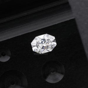 May include: A faceted, oval-cut diamond with brilliant white color. The gemstone is set against a dark, textured background. The image includes the text "OURO JEWELS" at the bottom.