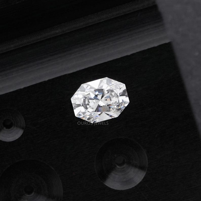 May include: A faceted, oval-cut diamond with brilliant white color. The gemstone is set against a dark, textured background. The image includes the text "OURO JEWELS" at the bottom.