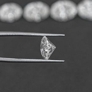 Pu&ograve; includere: Un diamante rotondo e trasparente tenuto in una pinza su uno sfondo nero.