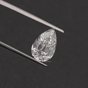 May include: A pear-shaped, clear gemstone with a slight pink hue. The gemstone is held by tweezers against a black background.