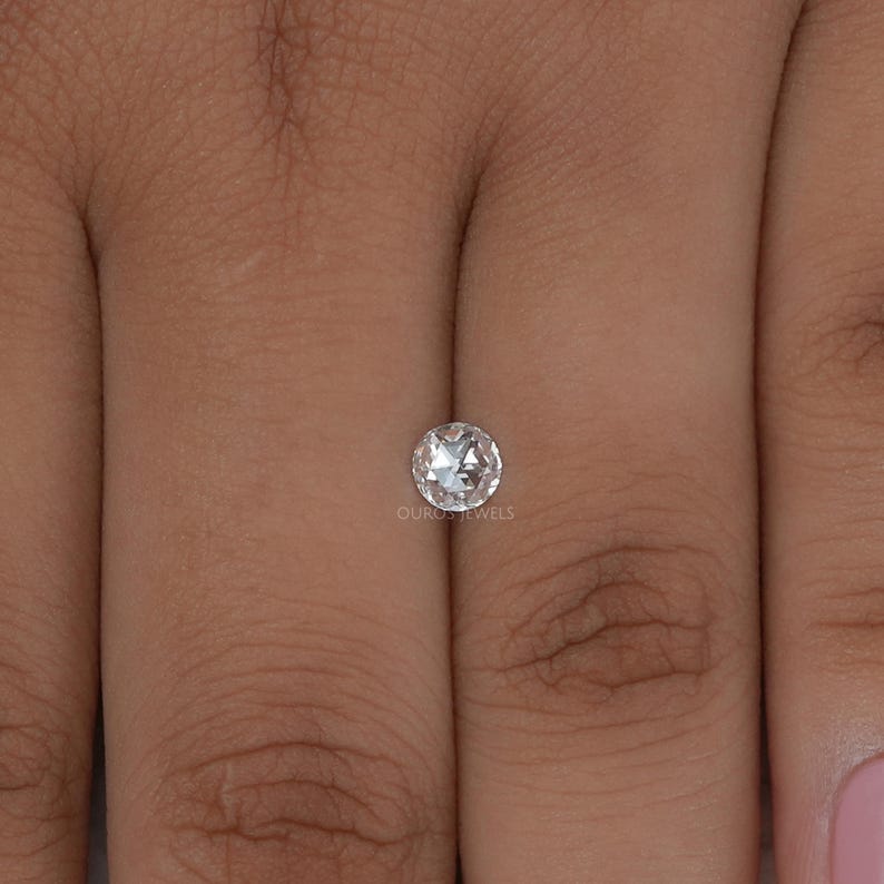 K&ouml;nnte beinhalten: Ein runder Diamant im Rosenschliff mit facettierter Oberfl&auml;che und klarem, farblosem Aussehen. Der Edelstein ist mittig platziert und tr&auml;gt den Text OUROS JEWELS in kleinen, wei&szlig;en Buchstaben direkt unter dem Stein.