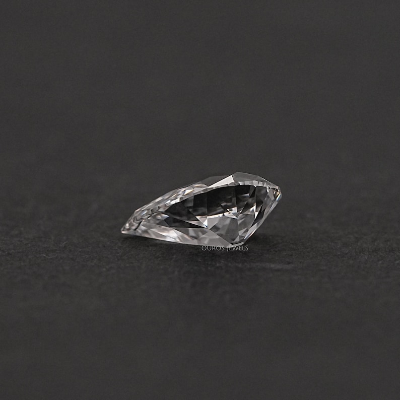 May include: A clear, pear-shaped gemstone with many facets. The gemstone is set against a black background.